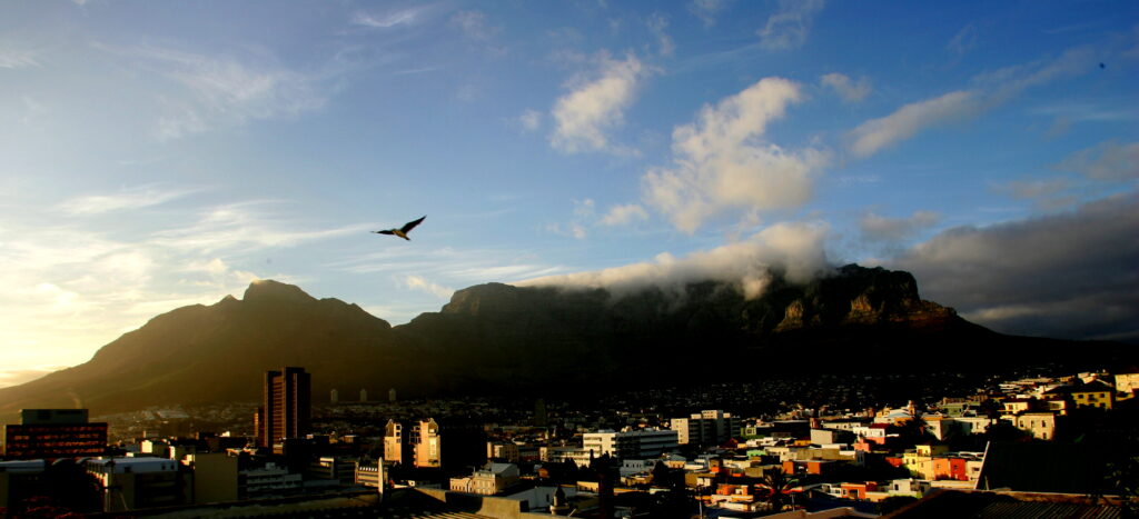 Nighttime view of Cape Town, with city lights below and Table Mountain silhouetted against a cloudy sky.