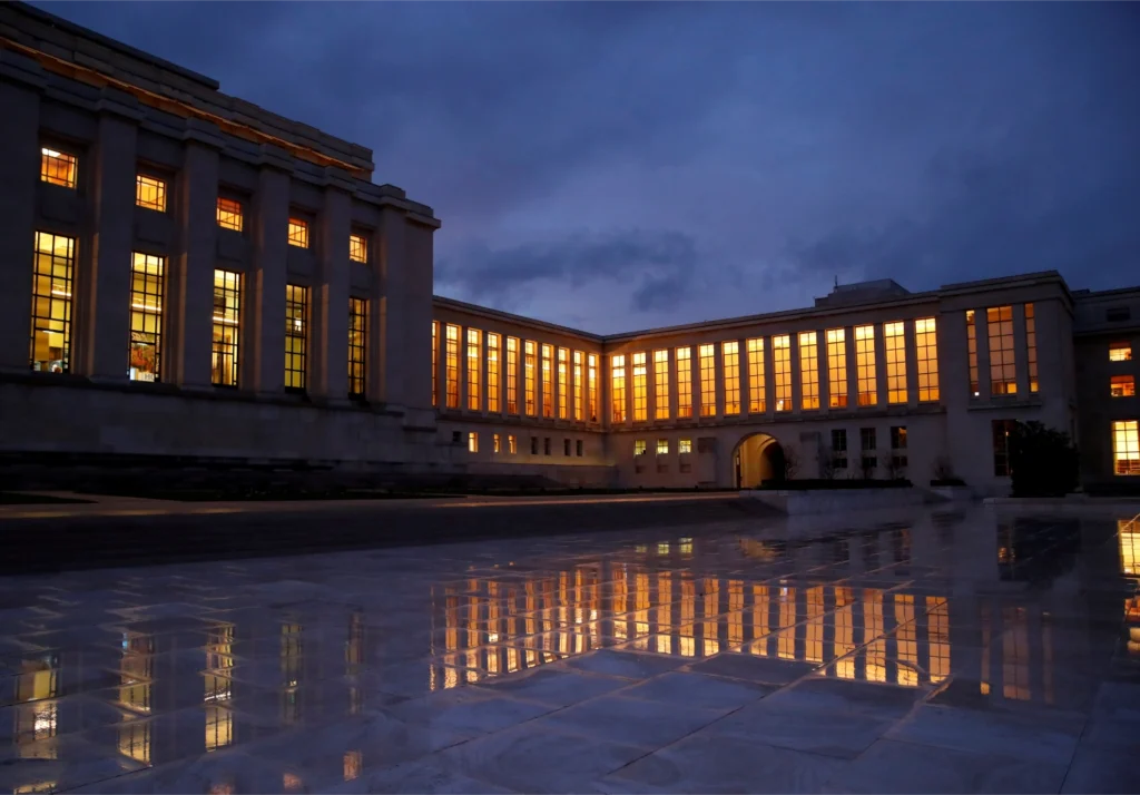 The United Nations building in Geneva at dusk, with illuminated windows reflected on a wet plaza in the foreground.